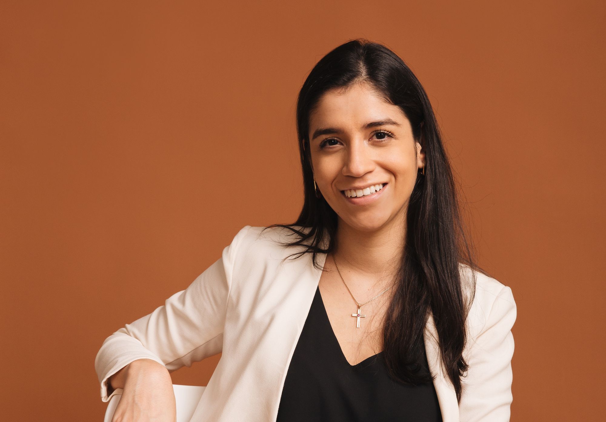 Portrait of a smiling Latina woman in a black shirt and cream blazer, sitting on a chair.